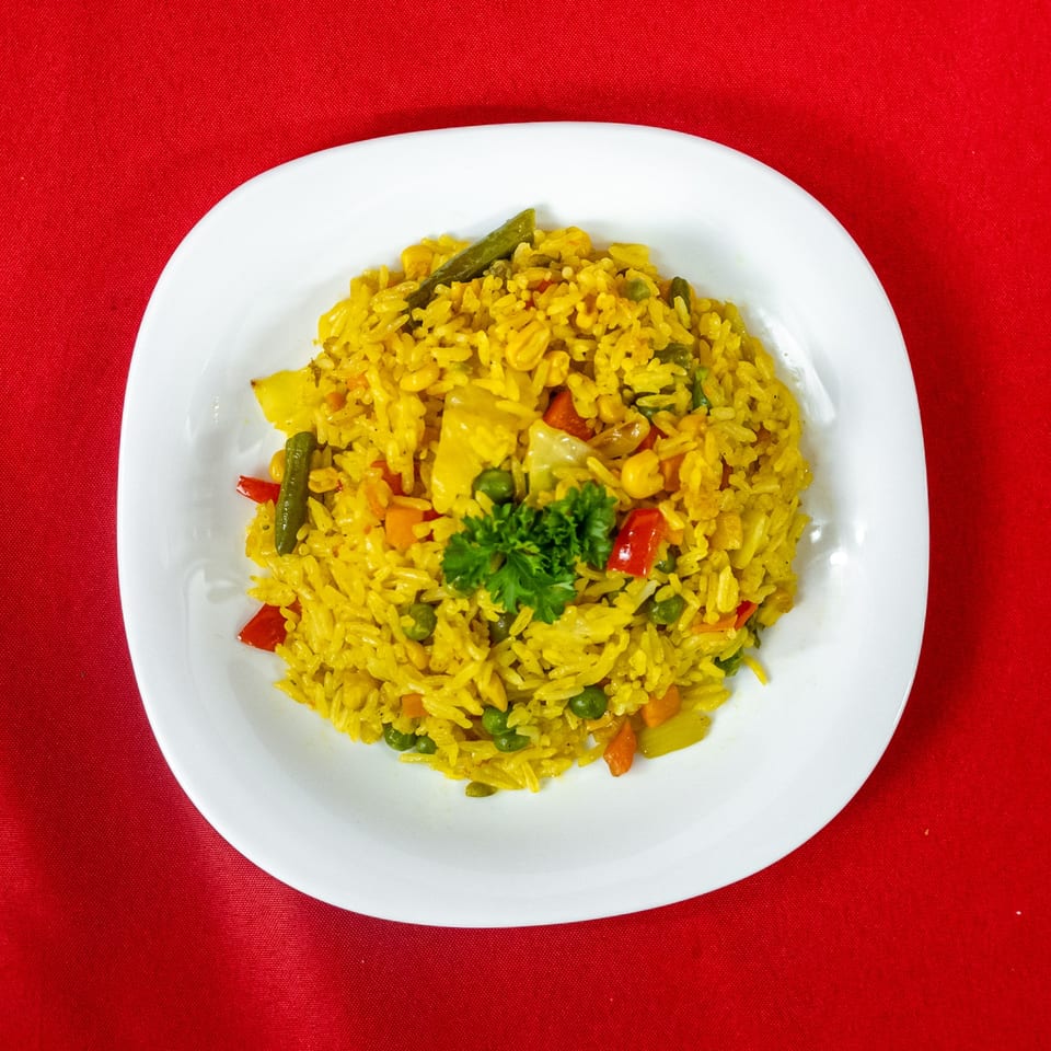 Yellow rice pilaf with mixed vegetables and parsley on a white plate against a red background.