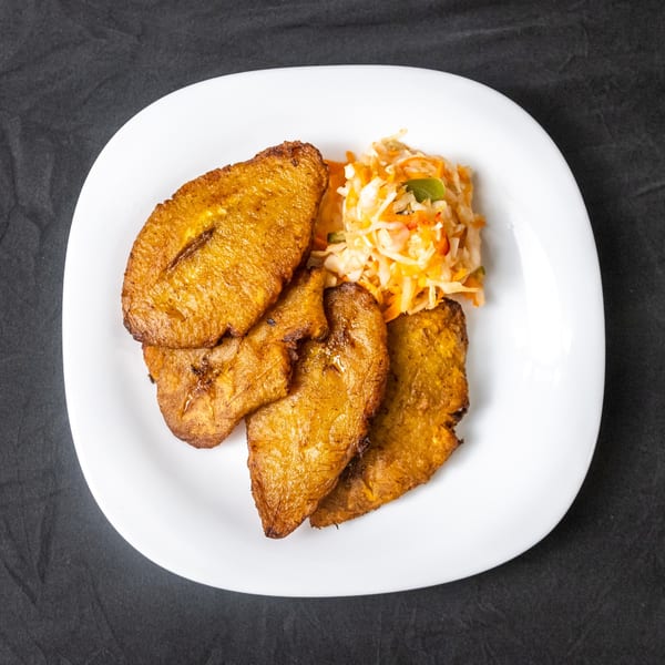 A white plate with golden-brown fried plantains and a side of coleslaw on a dark background.