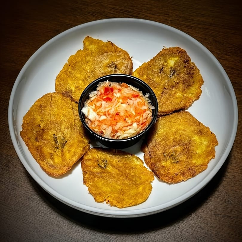 Five golden-brown fried plantains surround a small black bowl of pickled cabbage and peppers.
